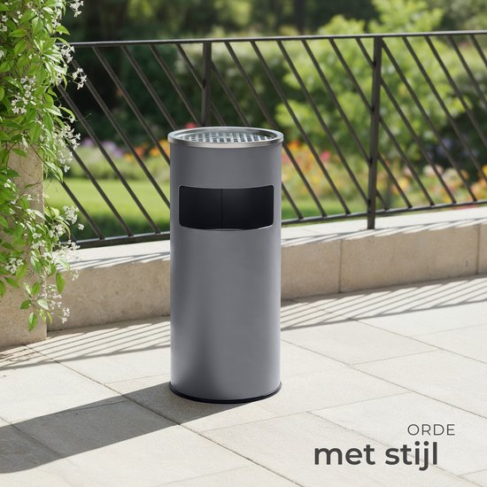Grijze staande asbak op een betegeld terras, met groene beplanting op de voorgrond en een hekwerk op de achtergrond.