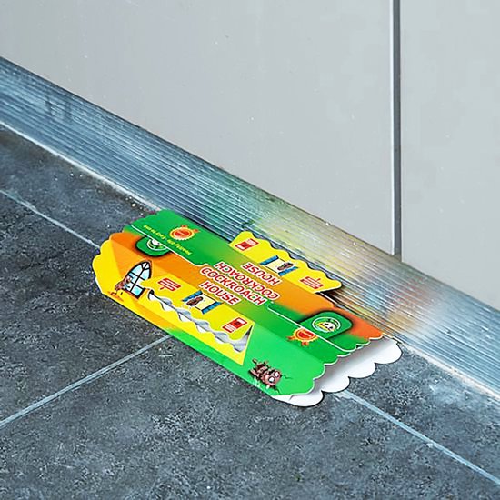 Close-up van een kokervormige kakkerlakkenval, met de tekst 'Cockroach House' en afbeeldingen van huizen en kakkerlakken, op een lichtgekleurde achtergrond.