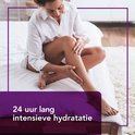 Vrouw met een witte blouse op een bed, brengt crème aan op haar benen. Tekst: 24 uur lang intensieve hydratatie.