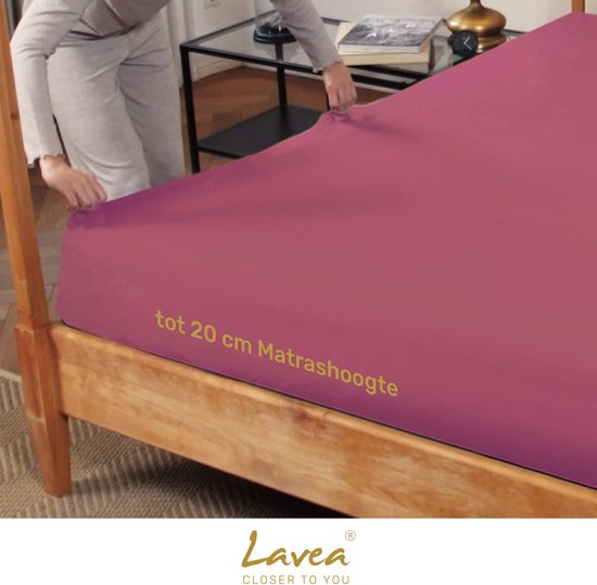 Een vrouw legt een koraalkleurig hoeslaken op een houten bed, geschikt voor matrassen tot 20 cm.
