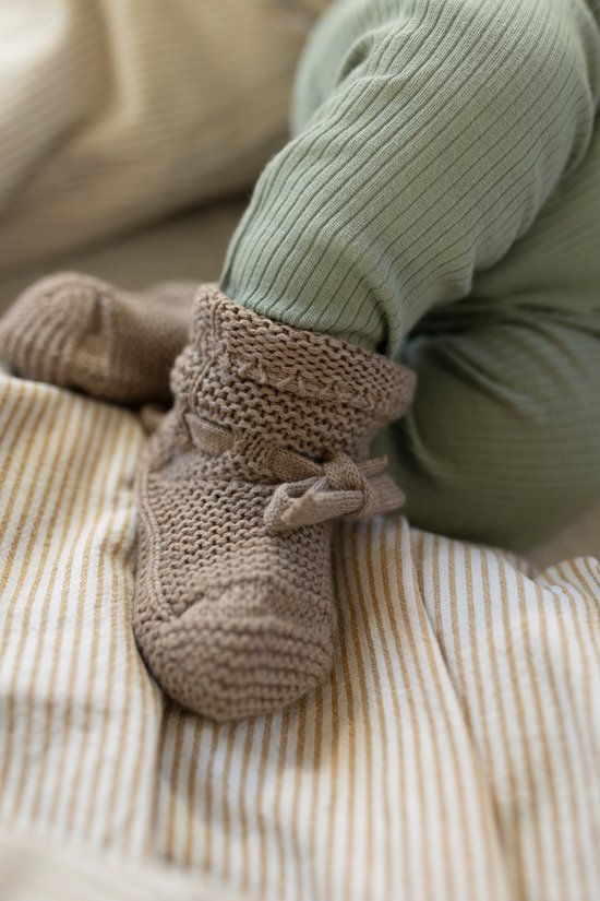 Baby's voetjes in gebreide taupe slofjes met strikjes, over een lichtgroene broek.