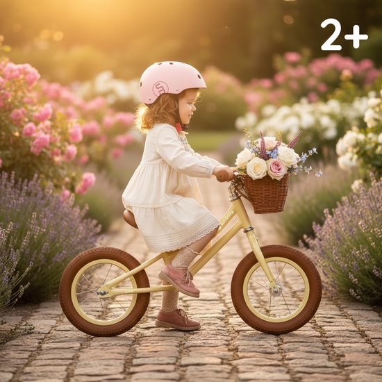 Meisje met roze helm en jurk zit op gele loopfiets met bloemenmandje in een tuin met roze en lavendelbloemen.