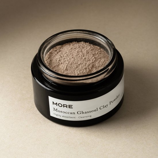 Glazen pot met zwarte deksel, gevuld met fijne, bruingrijze Marokkaanse Ghassoul kleipoeder. Zwart label met MORE en 'Moroccan Ghassoul Clay Powder'.