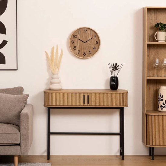 Ronde beige houten klok met zwarte cijfers, hangend boven een dressoir met ribbels.