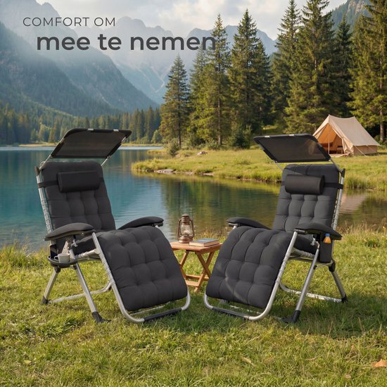 Twee zwarte, opklapbare ligstoelen met kussens, zonneschermen en bijzettafels, bij een meer met bergen en bomen op de achtergrond.