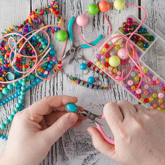 Handen maken een armband met een roze tang en diverse kleurrijke kralen, garens en sieraden op een witte achtergrond.