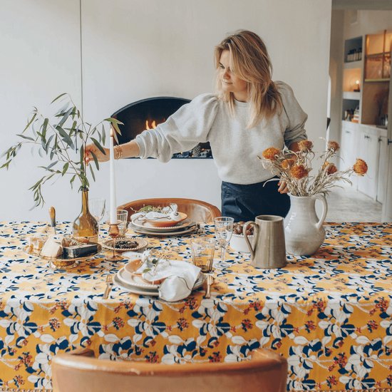 Vrouw legt een kaars aan een gedekte tafel met een geel bloemenkleed.