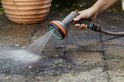 Een hand gebruikt een grijze Gardena sproeikop met oranje details om een stenen ondergrond schoon te spuiten.