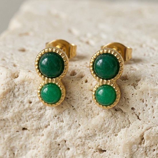 Gouden oorknopjes met twee ronde groene stenen op een stenen ondergrond.