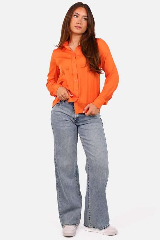 Model draagt een oranje blouse met kraag in glanzende stof en wijde lichtblauwe jeans.