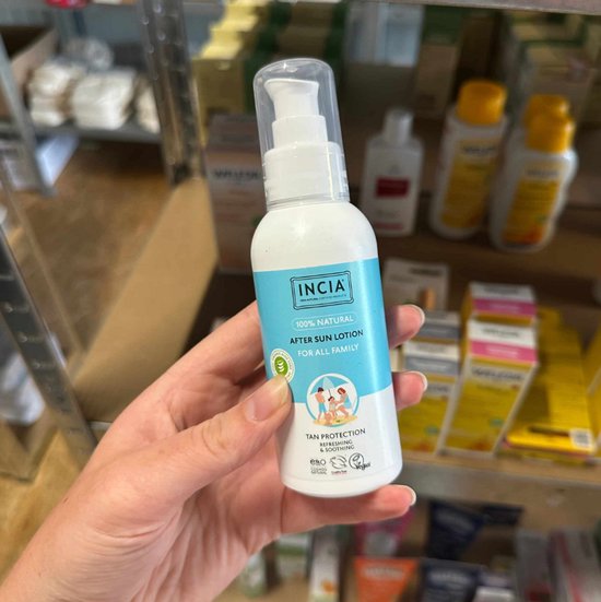 Witte fles met lichtblauwe label, vastgehouden door een hand. 'INCIA 100% NATURAL AFTER SUN LOTION FOR ALL FAMILY' en tekening van een gezin op het strand.
