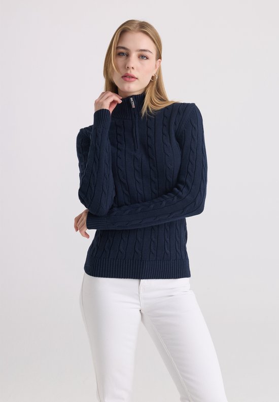 Model met donkerblauwe kabeltrui met ritskraag en witte broek.