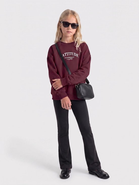 Meisje met zonnebril in bordeauxrode trui met tekst, zwarte broek en tas.