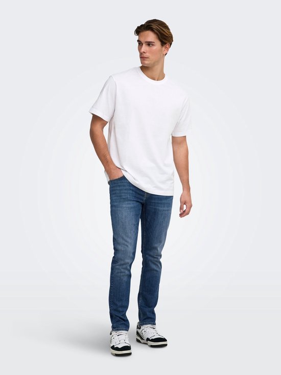 Model in een wit T-shirt en donkerblauwe slim-fit jeans met één hand in de broekzak, staand op een lichte achtergrond.