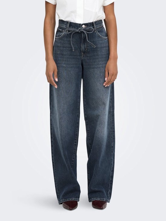 Model draagt een donkerblauwe spijkerbroek met een strikceintuur en wijde pijpen. De broek heeft een casual, stoere uitstraling.