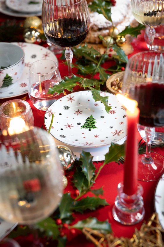 Feestelijke kersttafel gedekt met wit porseleinen servies, versierd met groene kerstbomen en rode sneeuwvlokken.