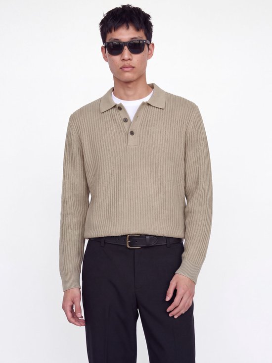 Close-up van model in beige gebreide polo met zwarte broek en riem.
