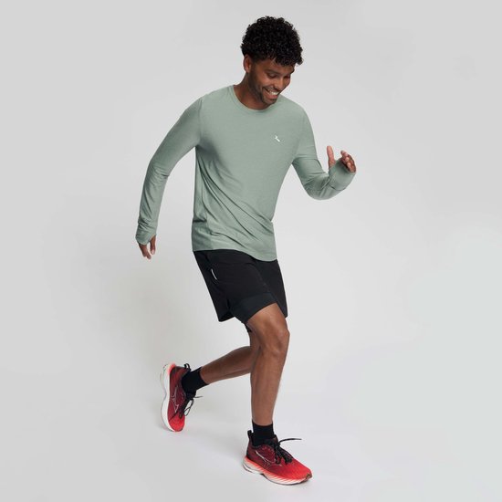 Man in beweging draagt een groen Rogelli hardloopshirt met lange mouwen, zwart sportbroekje en rode hardloopschoenen.