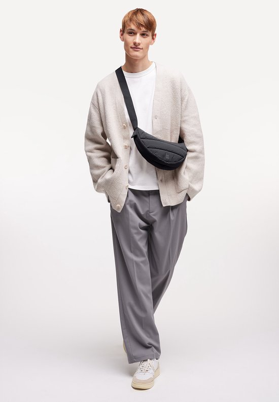 Man draagt een zwarte, gewatteerde crossbodytas over een beige vest en grijze broek.