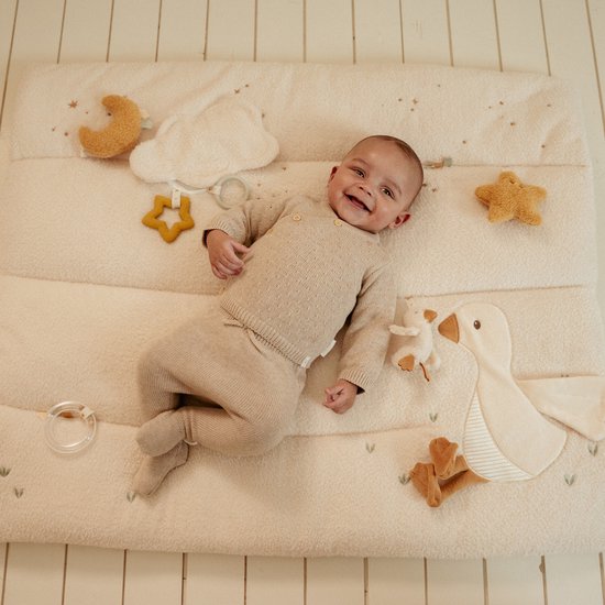 Baby ligt op een witte boxmat met maan, sterren, wolk en gans. Baby draagt beige trui.