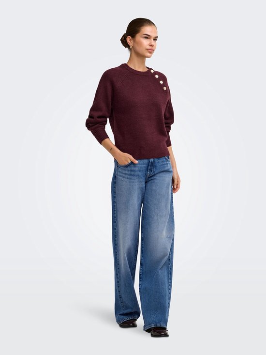 Model draagt een wijnrode gebreide trui met gouden knopen op de schouder en wijde jeans.