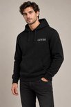 Man met donker haar en een baardje, gekleed in een zwarte hoodie met COVAR-logo en zwarte jeans.