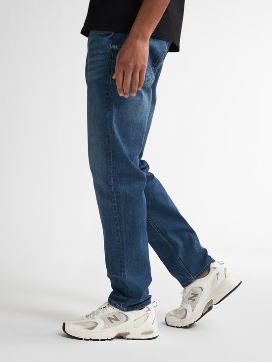 Model draagt een blauwe Petrol Industries jeans met regular tapered fit en witte New Balance sneakers.