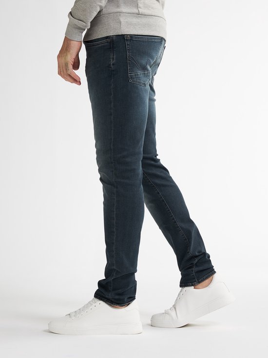 Model draagt een donkerblauwe slim fit jeans met vintage wassing, gecombineerd met een grijze trui en witte sneakers.