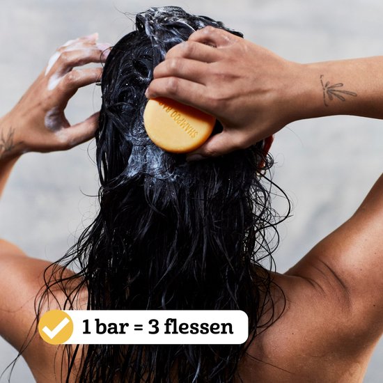 Een vrouw met donker haar wast haar hoofd onder de douche met een ronde, oranje shampoobar.