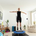 Een man in een zwart sportshirt en korte broek springt op een blauwe trampoline in een woonkamer.