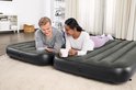 Man en vrouw liggen op twee aparte Bestway luchtbedden in een woonkamer. De man kijkt op zijn telefoon, de vrouw houdt een mok vast.