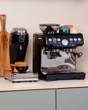 Zwarte koffiemolen en zilverkleurige espressomachine op een aanrecht, met gekleurde kopjes bovenop de machine.