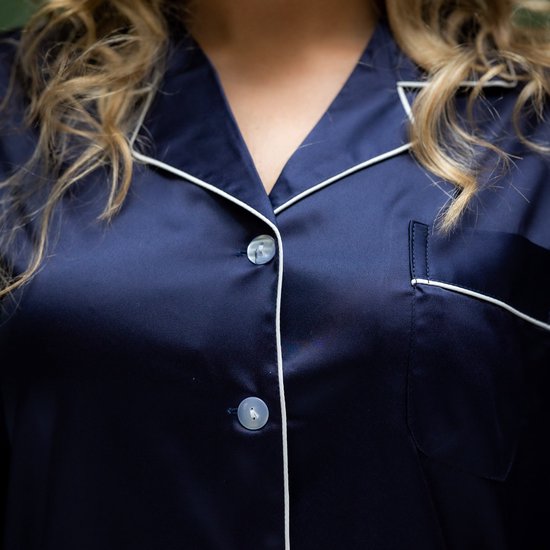 Close-up van de knoopsluiting en decolleté van een donkerblauwe satijnen pyjamajas met witte biesjes.
