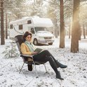 Vrouw leest in een stoel in een besneeuwde bosrijke omgeving, met een camper op de achtergrond. Ze draagt een gele jas en een groene sjaal. Het verwarmde zitkussen is zichtbaar in de stoel.