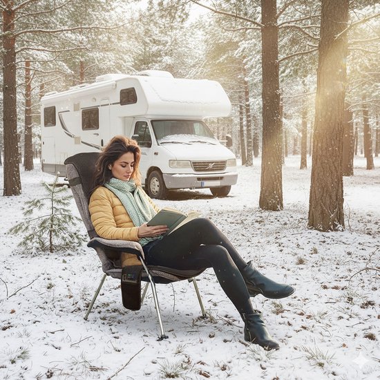 Vrouw leest in een stoel in een besneeuwde bosrijke omgeving, met een camper op de achtergrond. Ze draagt een gele jas en een groene sjaal. Het verwarmde zitkussen is zichtbaar in de stoel.