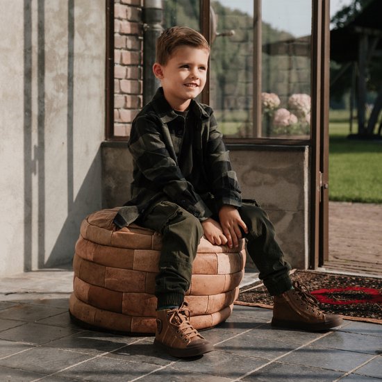 Jongen in geruite jas en bruine cargo broek zit op een bruine poef.