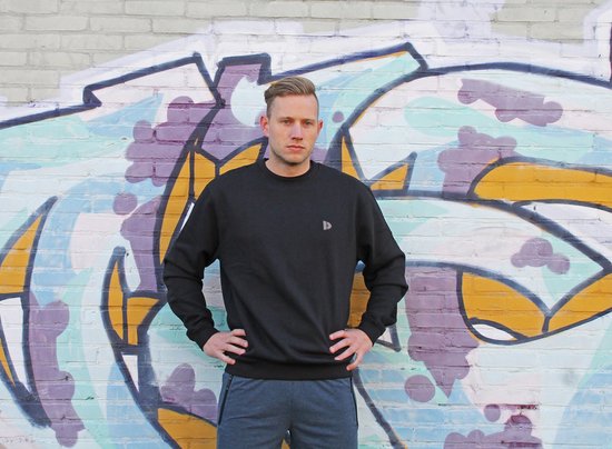 Man in zwarte Donnay fleece trui en grijze trainingsbroek voor een graffiti muur.