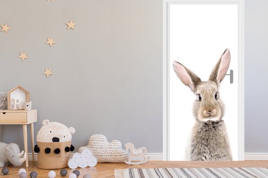 Een bruin, pluizig konijn kijkt nieuwsgierig door de deuropening in een kinderkamer. De deur is wit.