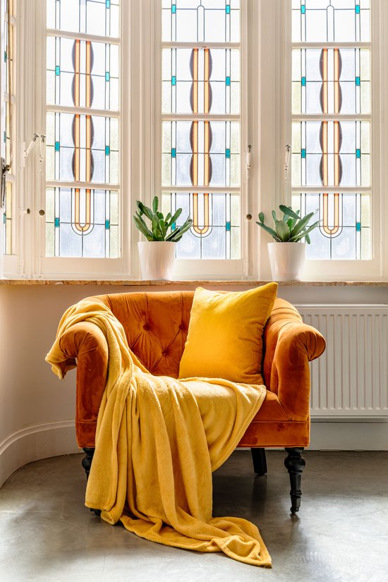 Een zachtgele, opgevouwen microvezeldeken ligt over een oranje fauteuil met een gele sierkussen.