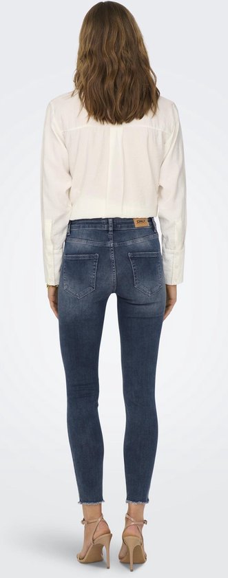 Vrouw van achteren gezien, in een donkerblauwe skinny jeans met rafelige zoom, met beige sandalen.