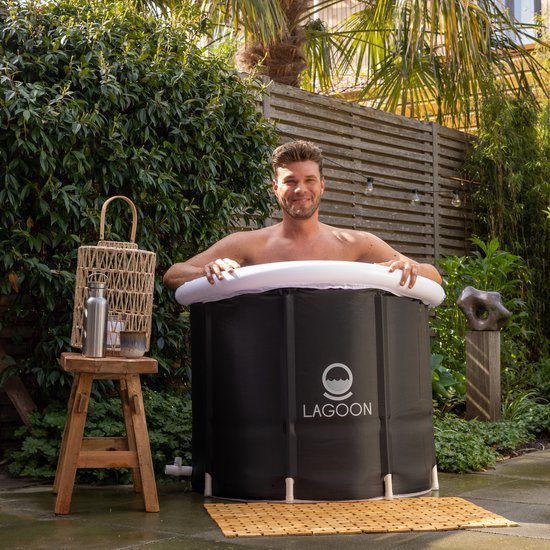 Man in een zwart, opvouwbaar bad met witte opblaasrand, in een tuin met houten schutting en planten.
