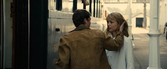 Een man met een suède jas legt een hand op de wang van een vrouw met blond haar, bij een trein.