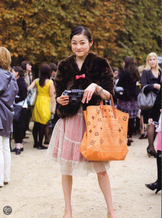 Vrouw met bruine bontjas, strikje, roze jurk, camera en oranje tas met LV-logo.