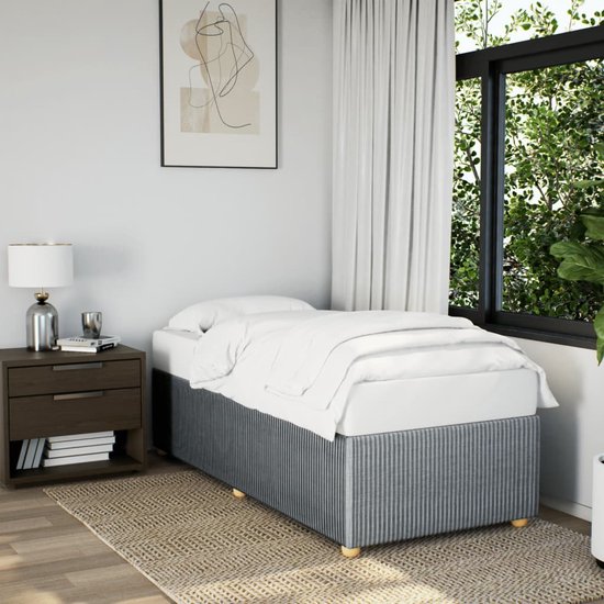 Intérieur de chambre avec sommier en textile gris clair, matelas avec draps blancs, table de chevet et fenêtre.