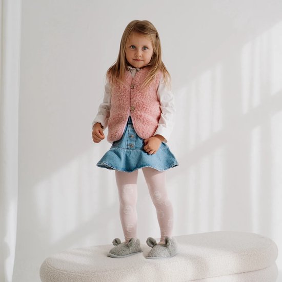 Meisje met blond haar, draagt een roze wollen gilet, wit overhemd, spijkerrokje, roze maillot en grijze muizensloffen.