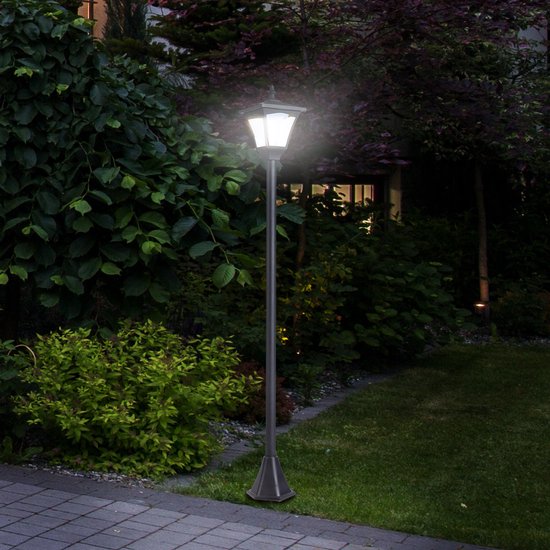 Verlichte zwarte lantaarnpaal in een weelderige tuin met struiken en gras.