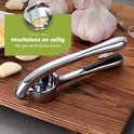 Zilverkleurige knoflookpers met zwarte handgrepen, op een houten plank met knoflookteentjes en hele bollen knoflook.