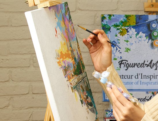 Gros plan sur des mains peignant une peinture par numéro. Paysage partiellement peint avec des ponts et des bâtiments, avec des pots de peinture au poignet.