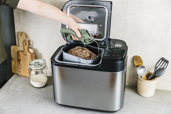 Zilverkleurige broodbakmachine met deurtje open, bakvorm eruit gehaald en een hand die brood erin doet.