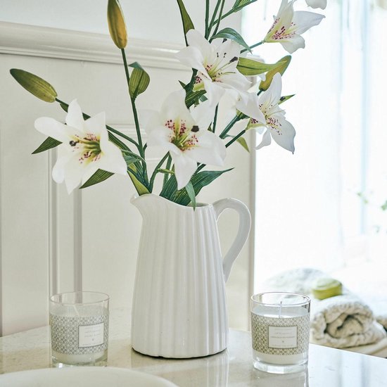 Witte karaf met verticale ribbels, met witte lelies in een vaas en twee geurkaarsen.